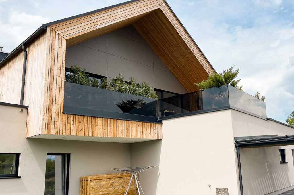 Modernes Haus mit Holzfassade und schrägem Dach, mit einem Glasbalkon mit Topfpflanzen und einem Wäscheständer darunter, vor einem teilweise bewölkten Himmel.