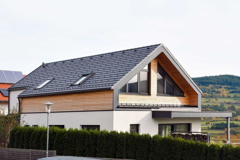 Modernes zweistöckiges Haus mit schwarzem Schrägdach, großen Glasfenstern und hellen Holzverkleidungen als Akzent. Grüne Hecken säumen das Grundstück und im Hintergrund ist eine hügelige Landschaft zu sehen.