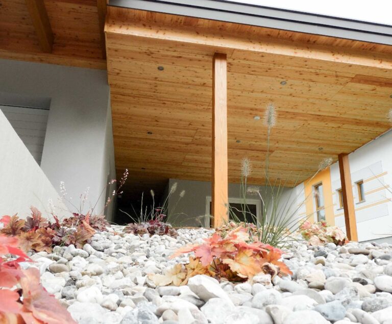Tiefblick auf ein modernes Gebäude mit einem hölzernen Überhang, der von Balken gestützt wird. Im Vordergrund sind dekorative Felsen und herbstlich gefärbte Pflanzen zu sehen, die zum Eingang hinführen.