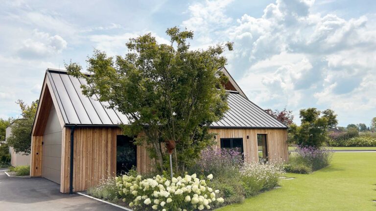Ein modernes Haus mit Holzverkleidung und einem Metalldach ist umgeben von grünem Gras, Bäumen und blühenden weißen Blumen unter einem teilweise bewölkten Himmel.
