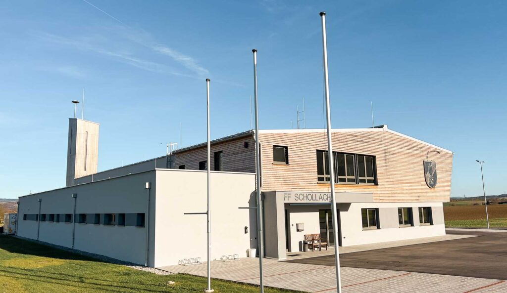 Ein modernes, zweistöckiges Feuerwehrhaus mit der Aufschrift "FF Schollach" und vier leeren Fahnenmasten davor steht auf einer gepflasterten Fläche neben einer Wiese unter blauem Himmel.