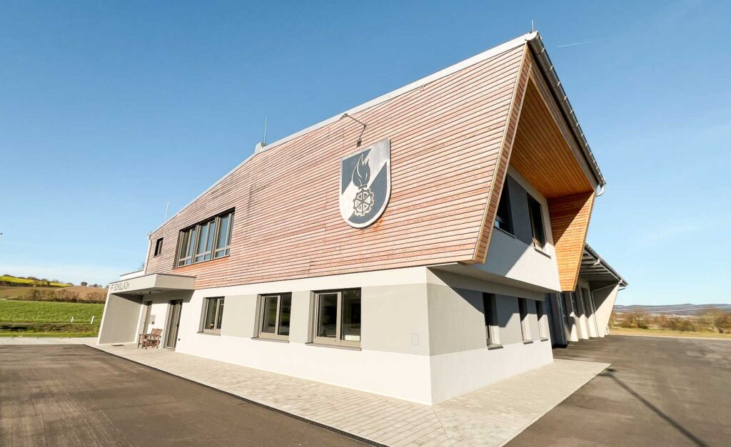 Modernes Feuerwehrgebäude mit hölzernem und weißem Äußeren, großen Fenstern und einem Feuerwehremblem an der Vorderwand; klarer blauer Himmel und grasbewachsene Felder im Hintergrund.