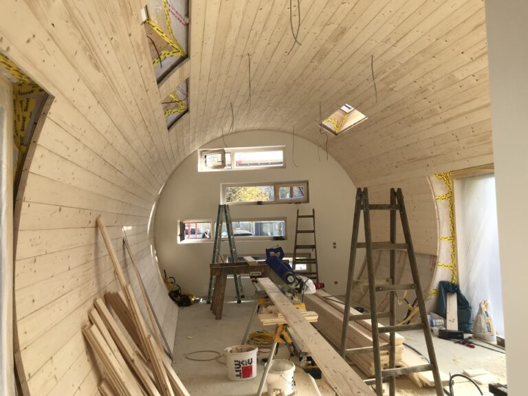 Das Innere eines teilweise gebauten, gewölbten Holzraums mit unfertigen Wänden und Decken, verstreuten Baumaterialien, Leitern, Holzbrettern und kleinen Fenstern, durch die Sonnenlicht einfällt.