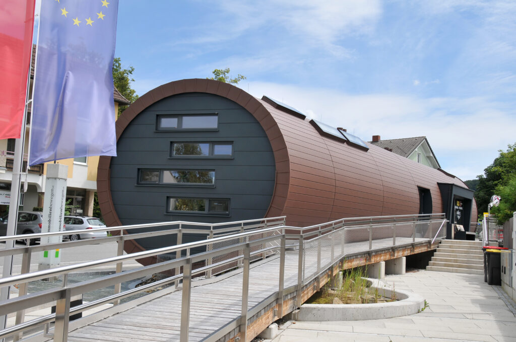 Ein modernes, zylindrisches Gebäude mit dunkler Verkleidung und rechteckigen Fenstern, zu dem man über eine schräge Rampe gelangt. Im Vordergrund sind die Flaggen der Europäischen Union und rote Fahnen zu sehen, und in der Nähe stehende Gebäude säumen die Straße.