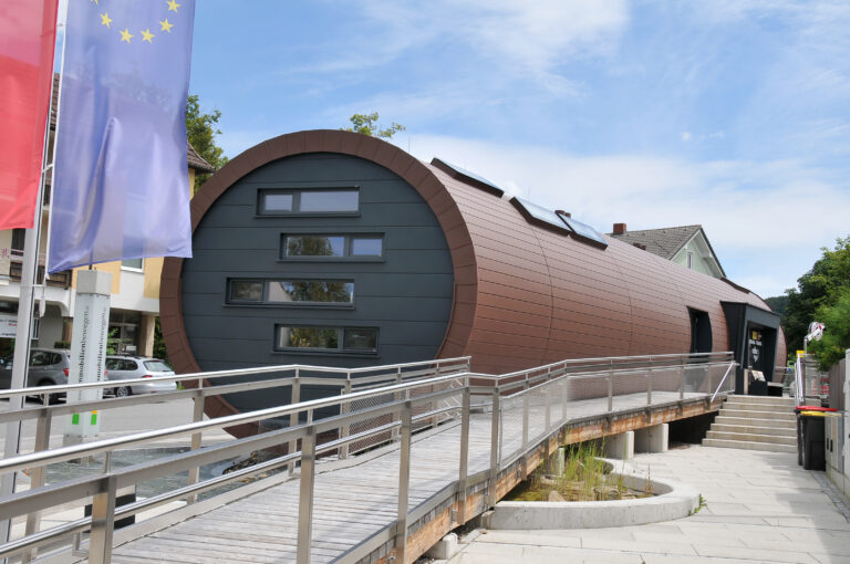 Ein modernes, zylindrisches Gebäude mit dunkler Verkleidung und rechteckigen Fenstern, zu dem man über eine schräge Rampe gelangt. Im Vordergrund sind die Flaggen der Europäischen Union und rote Fahnen zu sehen, und in der Nähe stehende Gebäude säumen die Straße.