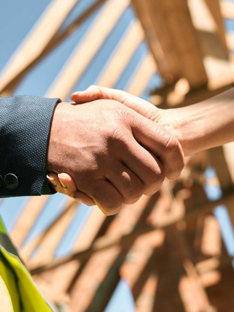 Zwei Personen schütteln sich im Freien die Hände, mit einem hölzernen Konstruktionsrahmen und blauem Himmel im Hintergrund. Eine Person trägt einen dunklen Anzug, die andere eine Warnweste.