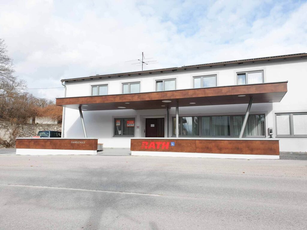 Ein modernes, zweistöckiges weißes Gebäude mit einer großen braunen Markise und einem Schild mit der Aufschrift "RATH" über dem Eingang, von einer ruhigen Straße aus gesehen an einem teilweise bewölkten Tag.