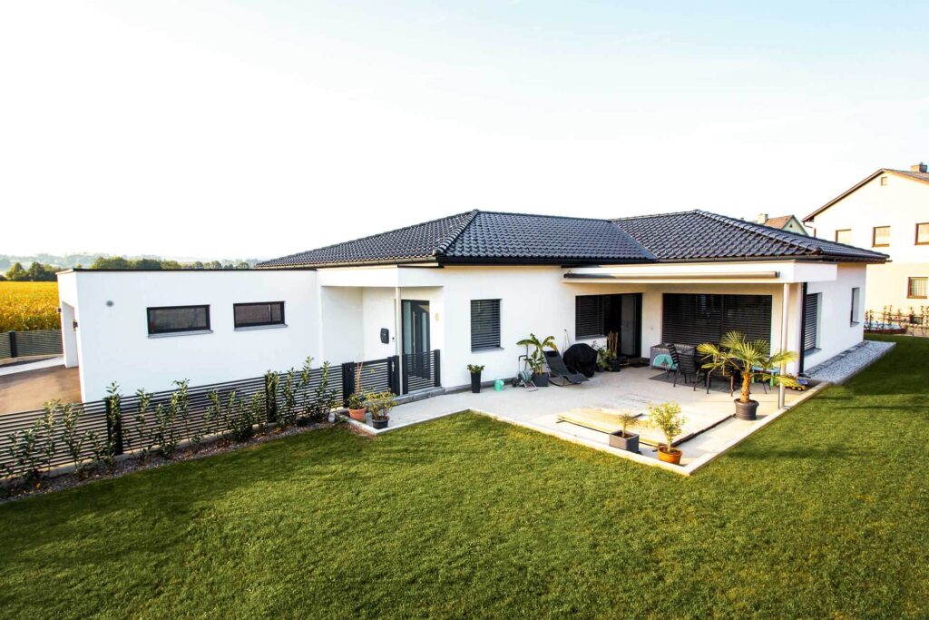 Modernes, einstöckiges Haus mit weißen Wänden und dunklem Ziegeldach. Es verfügt über eine geräumige Terrasse mit Gartenmöbeln und Topfpflanzen, die von einem gepflegten grünen Rasen umgeben und von niedrigen Hecken eingefasst ist.