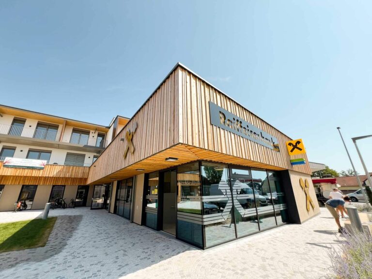 Ein modernes, holzgetäfeltes Gebäude mit großen Glasfenstern, auf denen das Logo der Raiffeisenbank prangt. Der Himmel ist klar, und eine Person geht in der Nähe auf einem gepflasterten Gehweg.