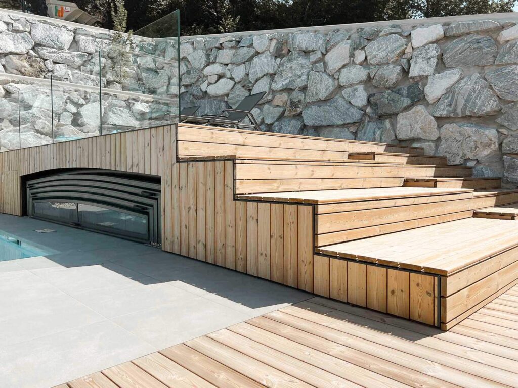 Moderne Außentreppe aus Holz an einer Steinmauer, mit Glasgeländer und einem kleinen Fenster oder einer Ablageöffnung unter der Treppe, die zu einem Poolbereich führt.