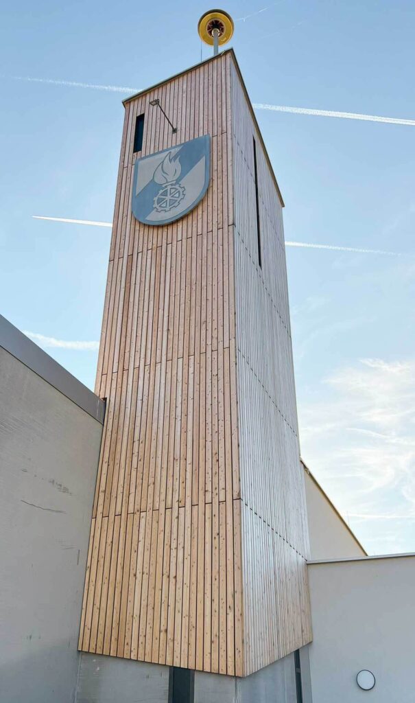 Ein hoher Holzturm mit vertikalen Paneelen erhebt sich vor einem blauen Himmel. Der Turm trägt ein schildförmiges Emblem mit einer Flamme und Symbolen. An der Spitze ist eine gelbe Sirene angebracht. Im Hintergrund ziehen Kondensstreifen über den Himmel.