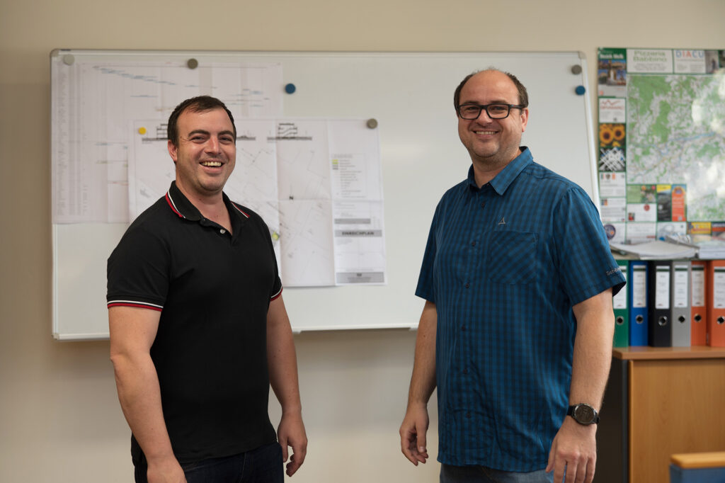 Zwei Männer stehen lächelnd vor einem Whiteboard, an das Papiere und Karten geheftet sind. Der eine trägt ein schwarzes Polohemd, der andere ein blau-kariertes Hemd. Im Hintergrund sind bunte Mappen und Karten zu sehen.