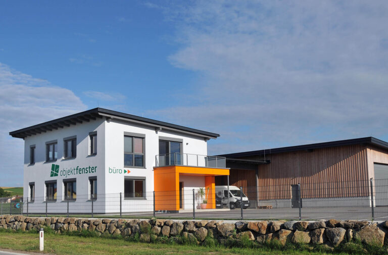 Ein modernes zweistöckiges weißes Bürogebäude mit einem orangefarbenen Eingang und dem Logo "objektfenster büro" steht neben einem großen Holzlager. Ein Lieferwagen ist in der Nähe geparkt, und eine Steinmauer und ein Zaun begrenzen das Grundstück.