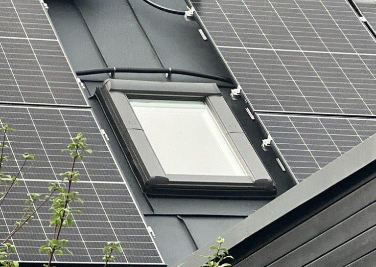 Nahaufnahme eines Dachfensters, das von Solarzellen umgeben ist, wobei in der unteren linken Ecke einige Äste zu sehen sind.